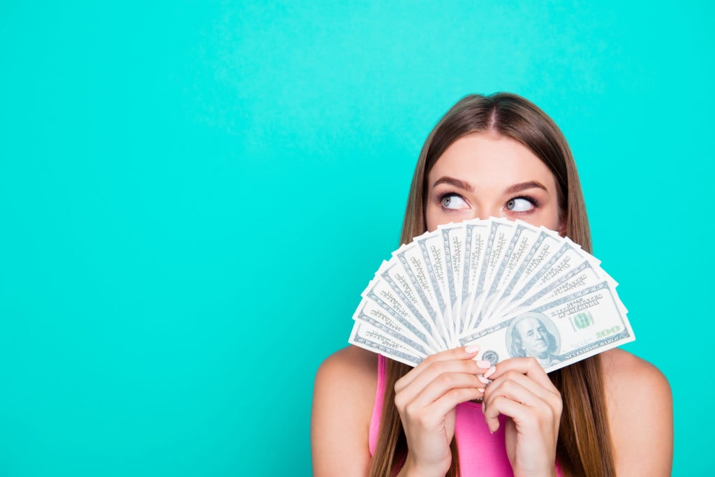 Attractive gorgeous young amazed girl wearing pink blouse, excited, covering face with dollar banknotes. Copy space. Isolated over bright vivid blue teal, turquoise background.