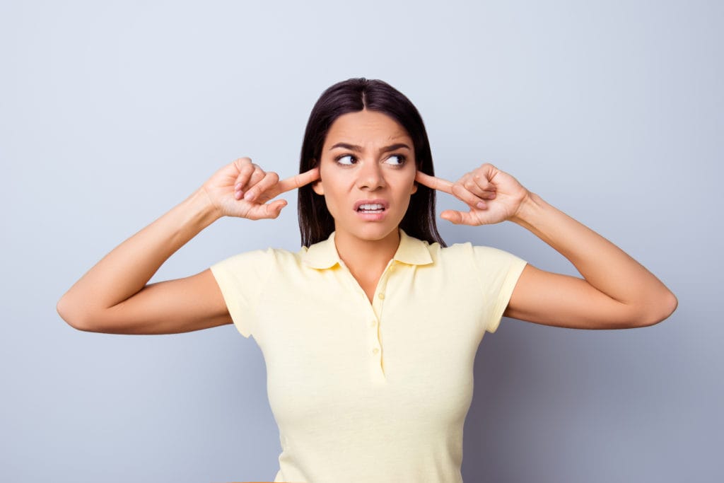 Close up of annoyed young latin American girl, closing her ears with fingers. She is looking angry, she is in a casual t-shirt on pure background.