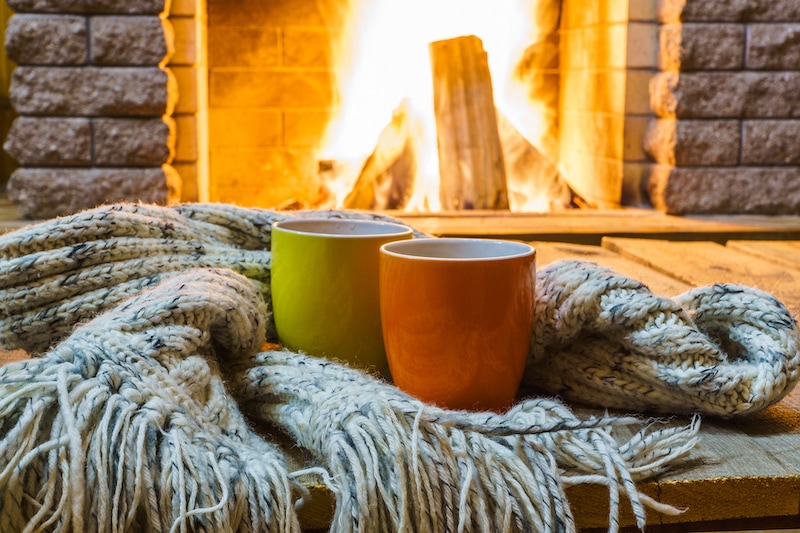 Two mugs for tea or coffee, woolen things near cozy fireplace, in country house, winter vacation, horizontal.