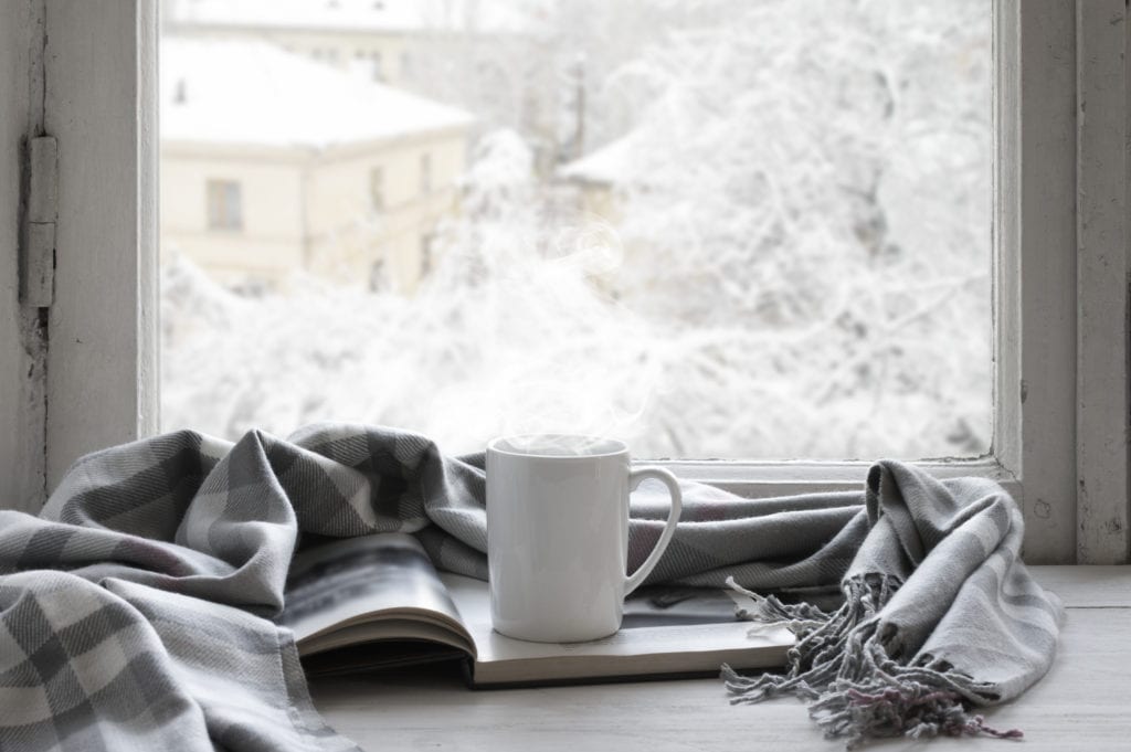 Cozy winter still life: mug of hot tea and opened book with warm plaid on vintage windowsill against snow landscape from outside.